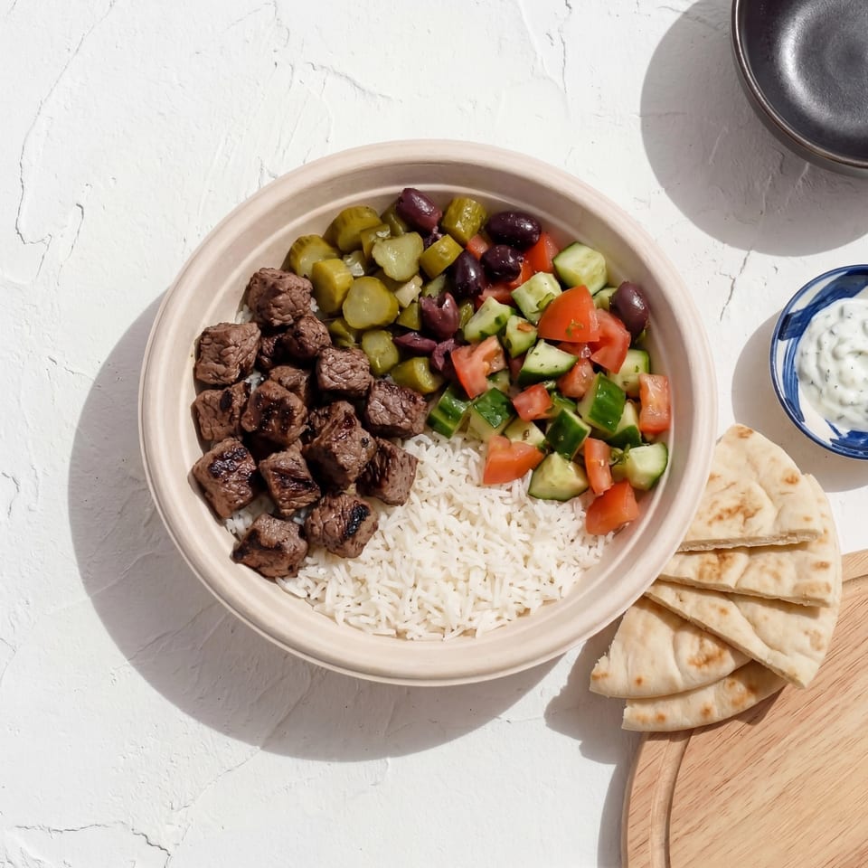 A beef and rice bowl with pickles, olives, and a cucumber tomato salad, with pita bread and tzatziki.